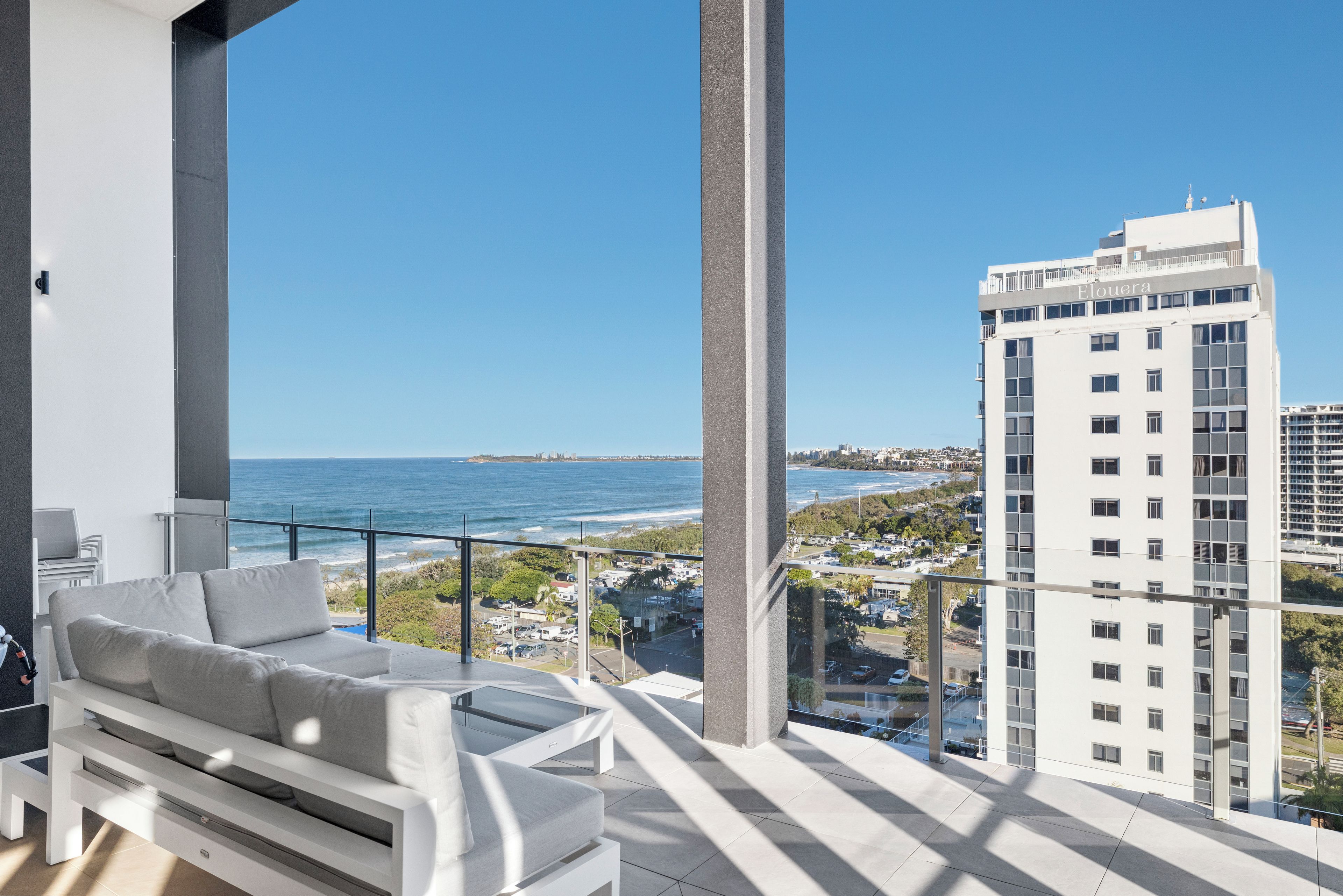 A balcony with a white sofa and a glass coffee table, overlooking a coastal cityscape with a clear view of the ocean and a tall building labeled 'Elouera'.