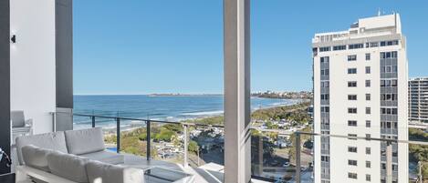 A balcony with a white sofa and a glass coffee table, overlooking a coastal cityscape with a clear view of the ocean and a tall building labeled 'Elouera'.