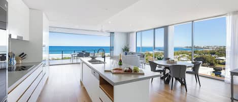 Une cuisine moderne dotée d’un grand îlot central, d’un coin repas offrant une vue sur l’océan et d’un vaste espace de vie avec de grandes fenêtres.