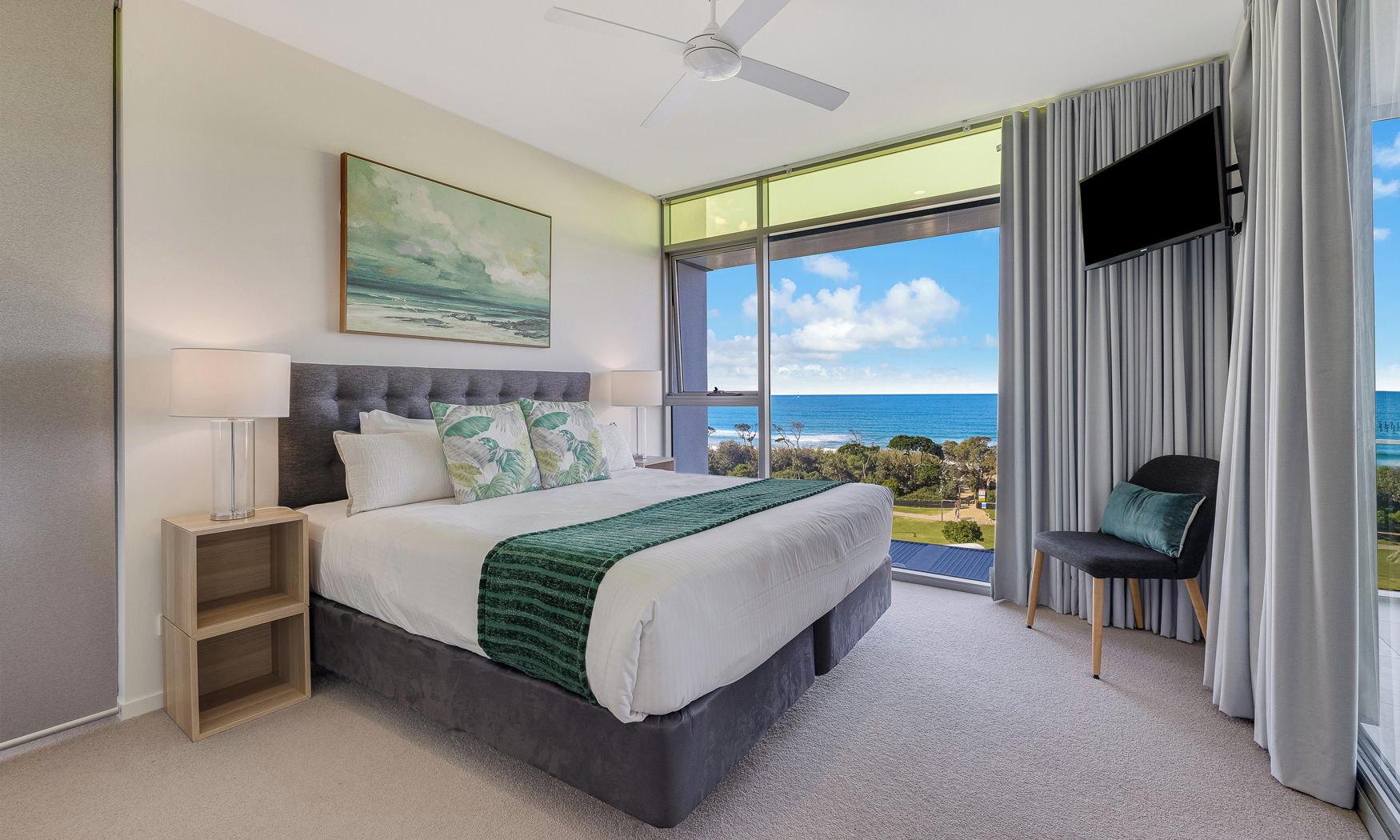 Quarto com cama, mesa de cabeceira, poltrona, televisão e uma grande janela com vista para o oceano.
