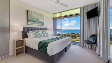 Quarto com cama, mesa de cabeceira, poltrona, televisão e uma grande janela com vista para o oceano.