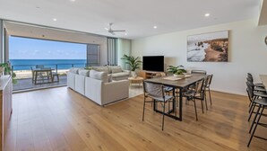 Una sala de estar moderna con un ventanal que da al mar, un televisor de pantalla plana y un comedor con mesa y sillas de madera.