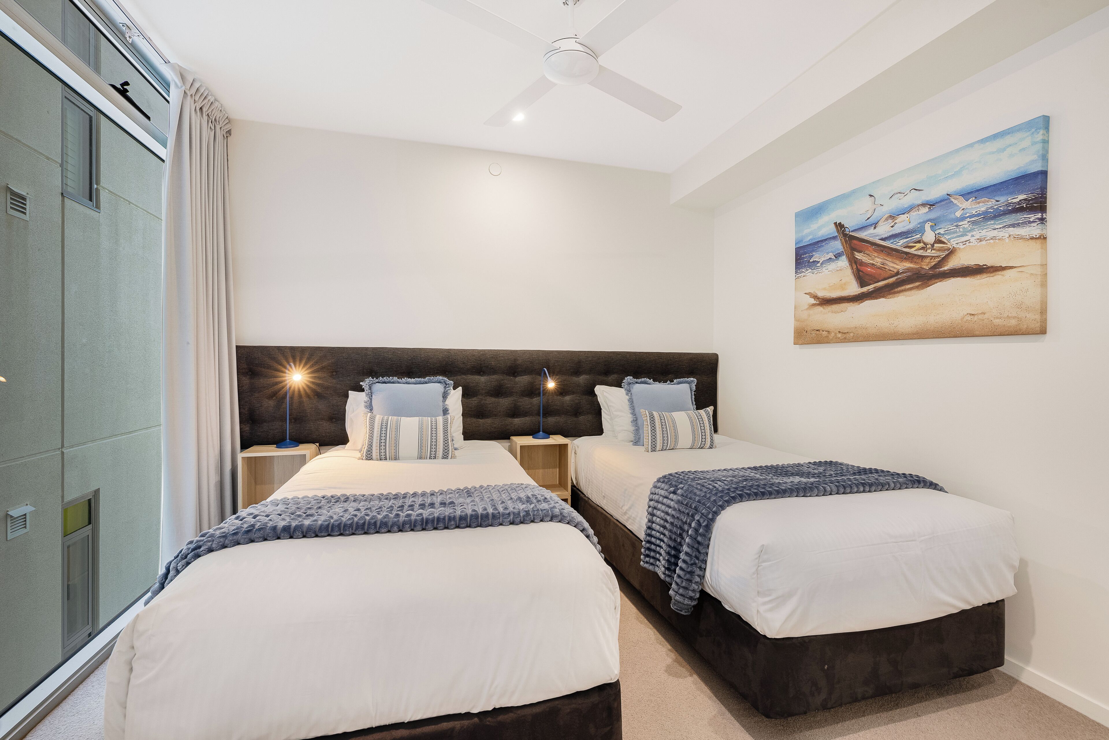 Habitación de hotel con dos camas, ventilador de techo, un cuadro de una playa y una ventana con cortinas.