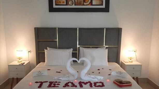 Une chambre d’hôtel avec un lit, deux tables de chevet, une œuvre d’art encadrée au-dessus du lit et une décoration en forme de cœur posée sur le lit.
