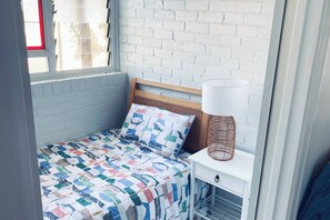 Una cama pequeña y bien hecha con un colcha colorida, un cabecero de madera, una mesita de noche blanca con una lámpara y una pared de ladrillo blanco.