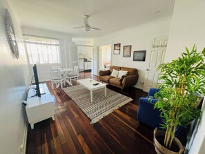 Een woonkamer met een bruine bank, een witte salontafel en een blauwe fauteuil. Er is een eethoek met witte stoelen en een witte tafel. In de achtergrond is een keuken zichtbaar.