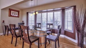 A dining area with a wooden table and chairs, a bar counter, and a view through large windows with blinds. - Moana Del Sol - Vineyard Estate + Resort Pool (Temecula)