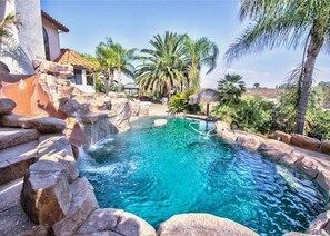 Una piscina cristalina rodeada de rocas y palmeras, con un edificio al fondo.
