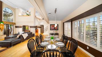 A spacious dining area with a long table set for six, surrounded by black chairs, in a modern interior with wooden flooring and large windows.