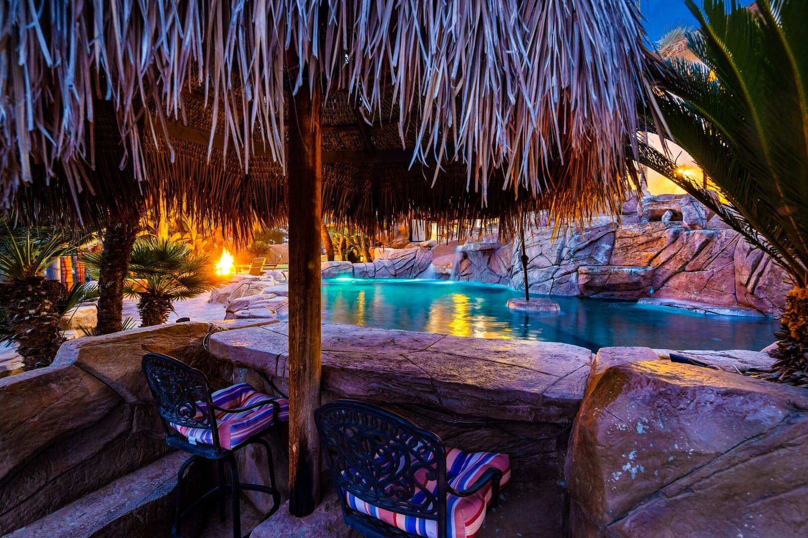 A pool area with a thatched roof, lounge chairs, and a waterfall.