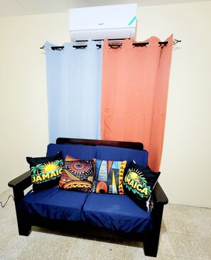 Ein Zimmer mit einem blauen Sofa, zwei Kissen mit tropischen Motiven und einer weißen Wandklimaanlage.