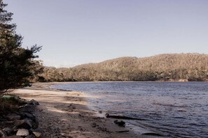 Blick auf einen See mit sandigem Strand, felsiger Uferlinie und bewaldeten Hügeln im Hintergrund.