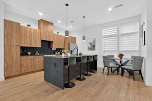 A modern kitchen with wooden cabinets, a central island with bar stools, and a dining area with a glass table and chairs. - Dallas Skyline Terrace Townhome (Dallas)
