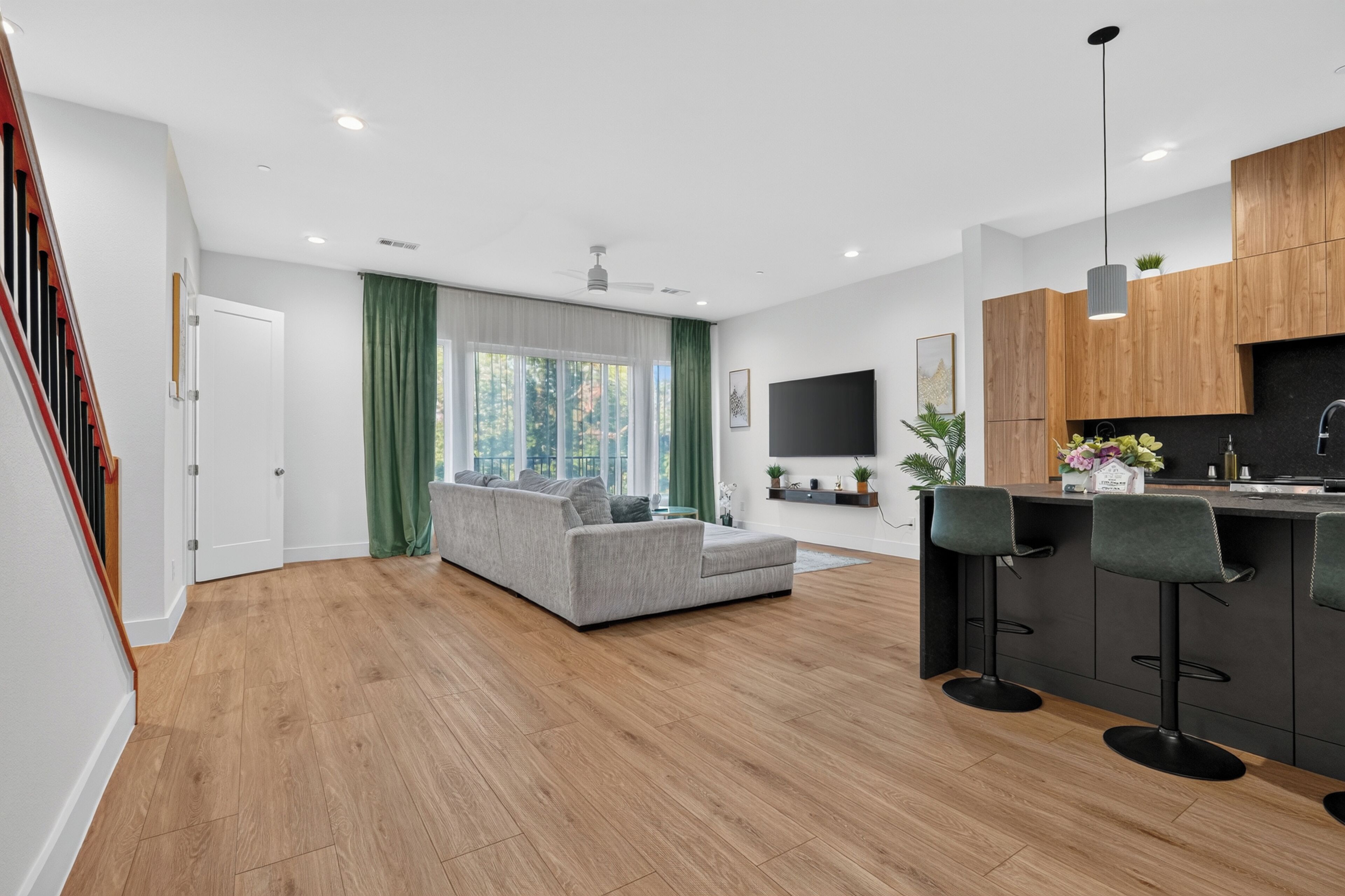 A modern living room with a grey sofa, wooden flooring, a green curtain, and a kitchen area with wooden cabinets and a bar.