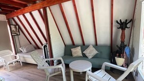 Une pièce avec un plafond en bois, un canapé vert orné de coussins à motifs, des chaises en plastique blanches et une table ronde.