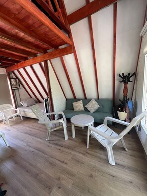 A room with a wooden ceiling, a green sofa with patterned cushions, white plastic chairs, and a round table.