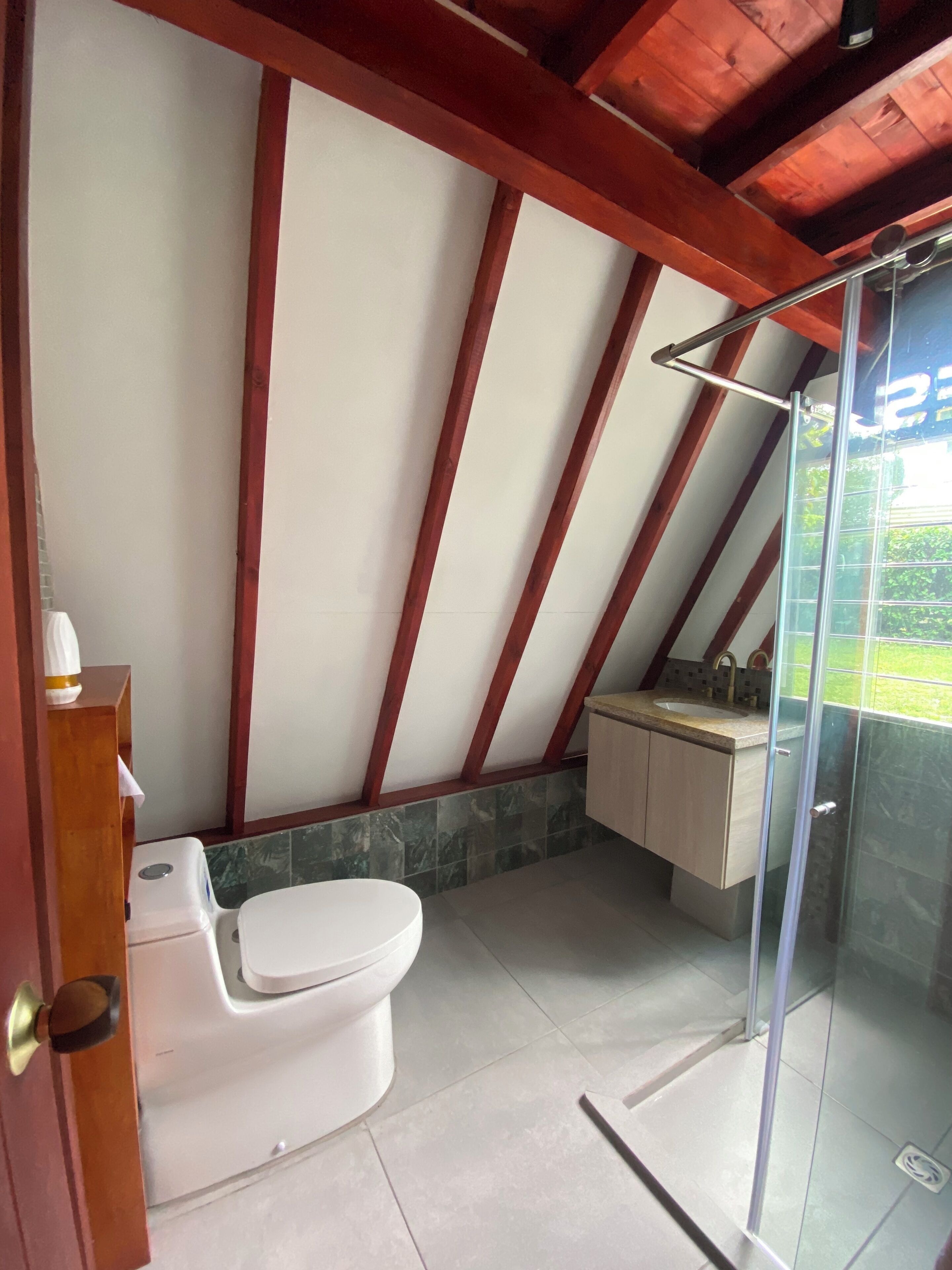 Baño con inodoro blanco, cabina de ducha de vidrio y estructura de techo de madera.