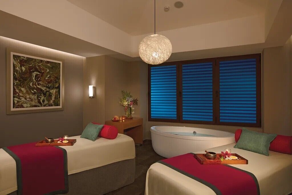 A spa room with two beds, a massage table, a bathtub, and a large window with blinds.