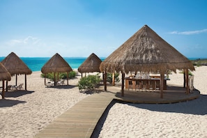 A beach with thatched-roof huts and a wooden deck leading to a bar area. - Luxury, Ocean view room at Dreams Playa Mujeres (Quintana Roo)