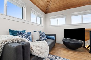 A modern living room with a grey sofa, blue patterned cushions, a fluffy white throw, and a black entertainment unit with a TV. - Blue Me Away | Family & Pet Friendly (Port St. Joe)
