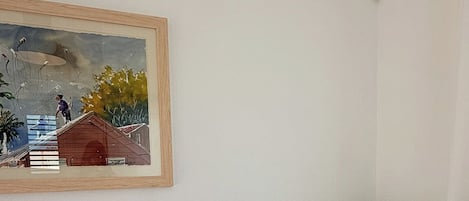 A room with a white wall, a framed painting, a white sofa with two pillows, and a wooden floor.