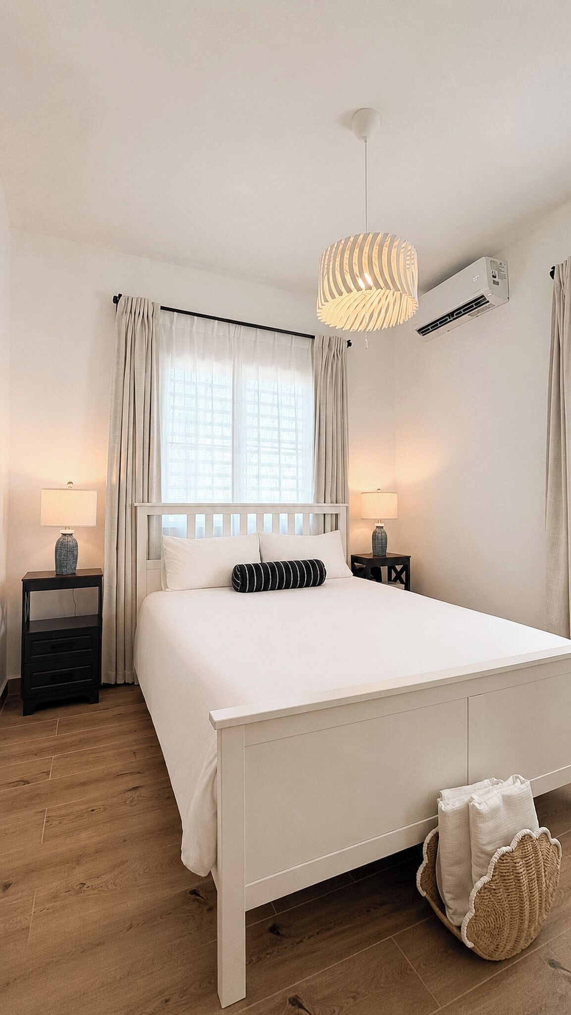 A hotel room with a white bed, two bedside tables with lamps, a hanging light fixture, an air conditioning unit, and a window with curtains.