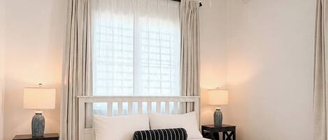 A hotel room with a white bed, two bedside tables with lamps, a hanging light fixture, an air conditioning unit, and a window with curtains.
