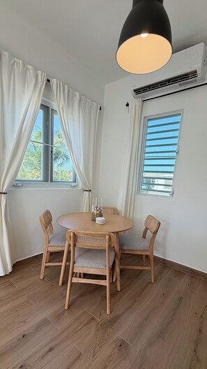 Una habitación con una mesa redonda de madera, dos sillas, una lámpara colgante, un equipo de aire acondicionado y ventanas con cortinas.