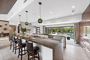 A modern kitchen with a central island, bar stools, pendant lighting, and a view of an outdoor pool area. - Sun-Drenched Coastal Haven with Private Pool (Newport Beach)