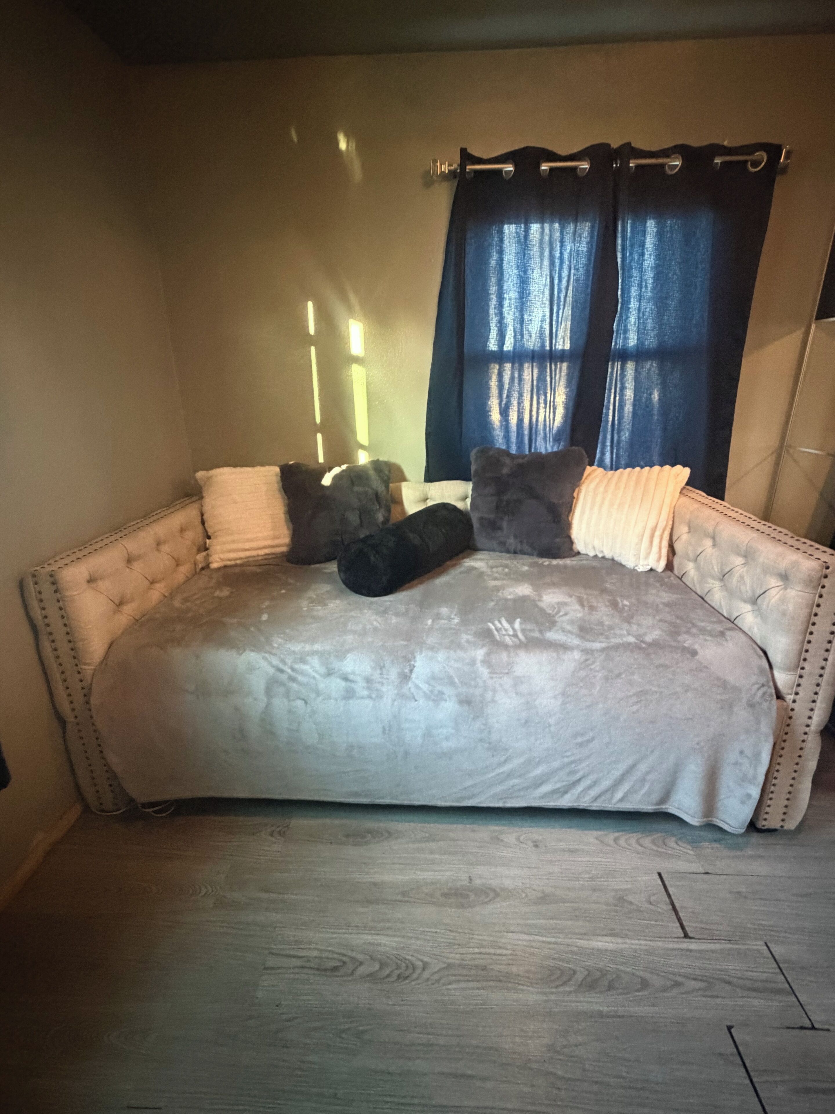 Una habitación con un sofá cama capitonado, una ventana con cortinas y suelo de madera.