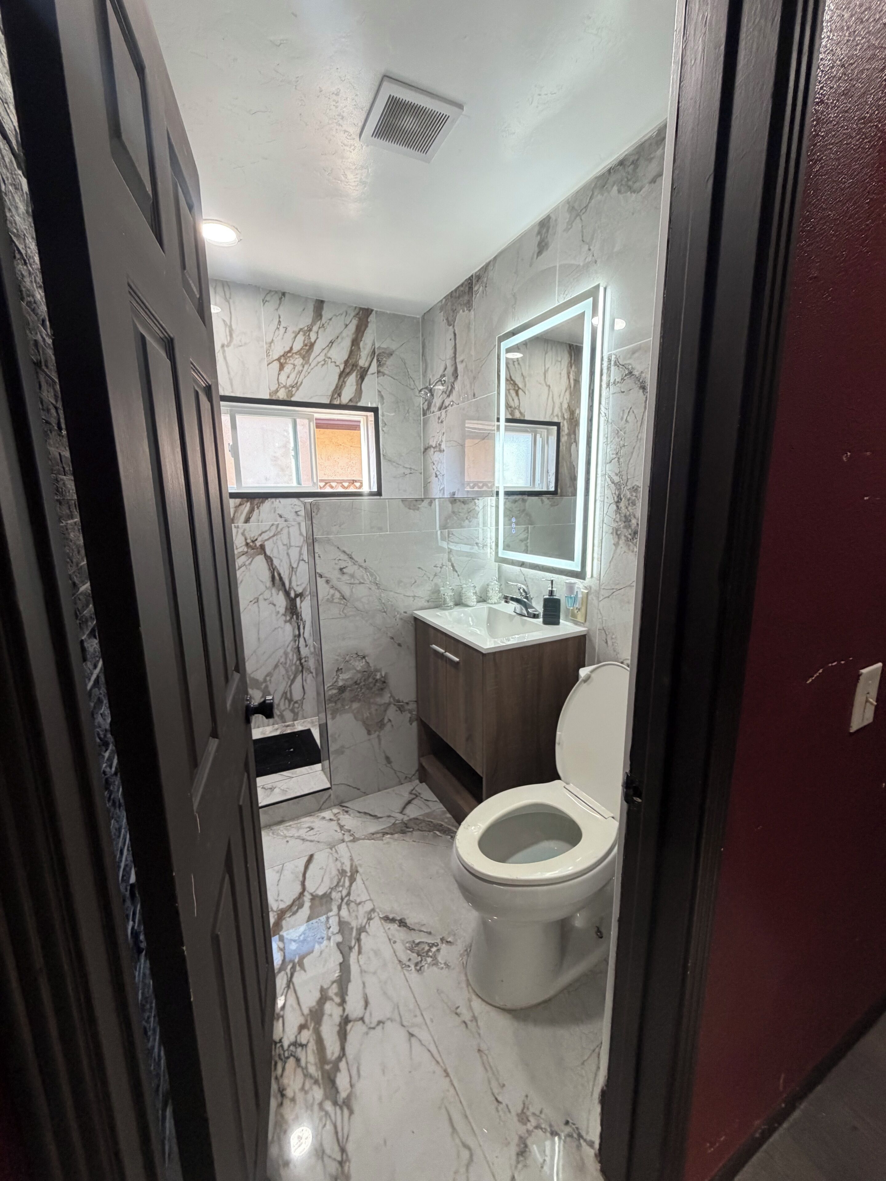 A bathroom with a marble floor, a white toilet, a wooden vanity, and a mirror with a light.