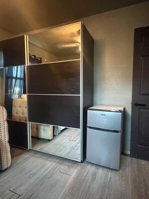 A compact room with a wardrobe, a small refrigerator, and a bed with a headboard.