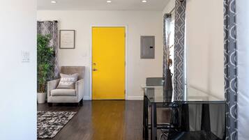 A room with a yellow door, a glass table, a chair, a plant, and a framed sign on the wall.