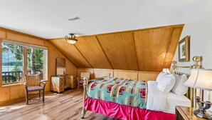 Ein Schlafzimmer mit Holzboden, einem Bett mit gemusterten Betttextilien, einem Nachttisch mit Lampe und einem Fenster mit Blick auf Bäume.