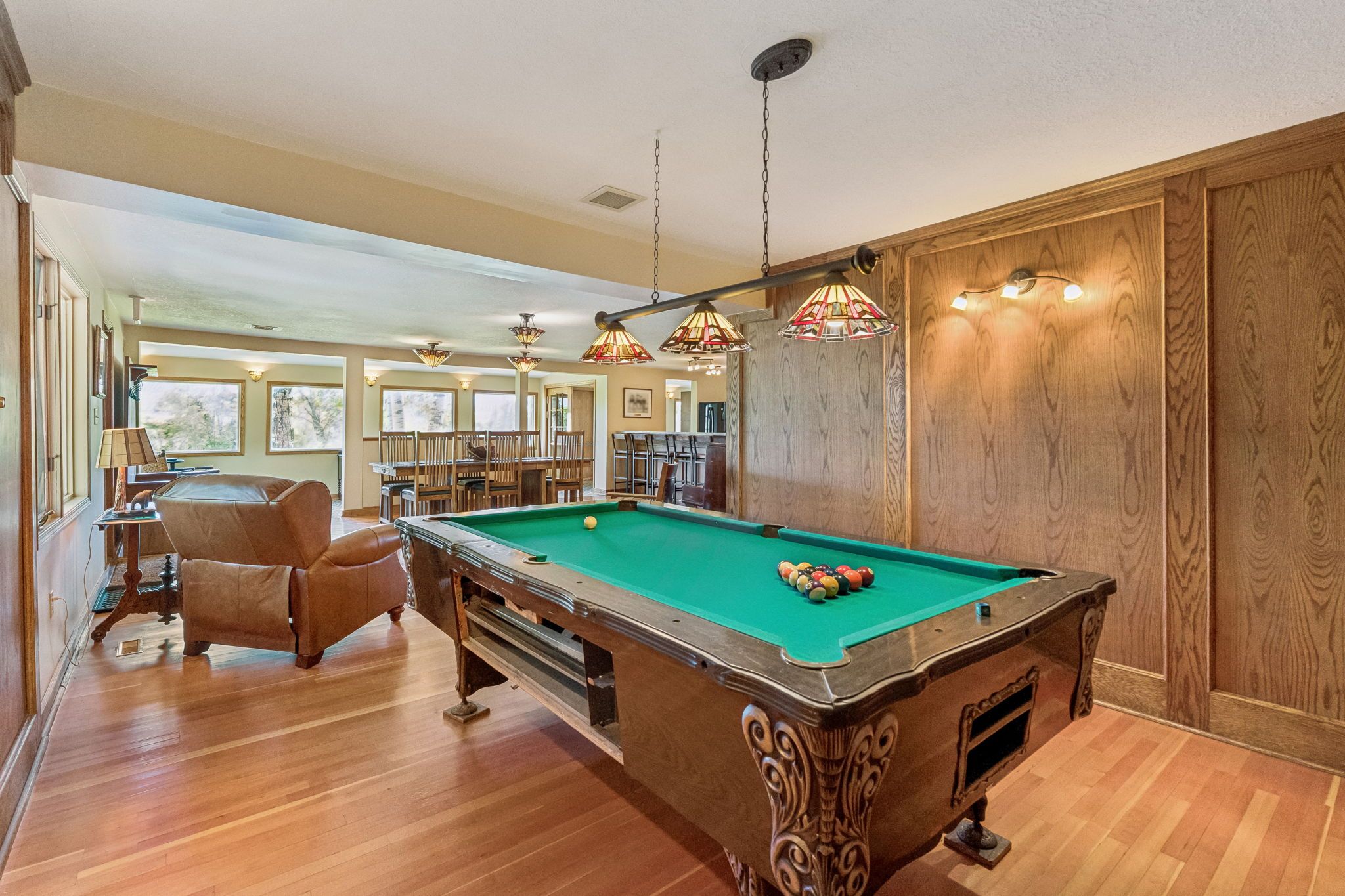 A pool table in a room with wooden flooring, a pool cue, and a chair.