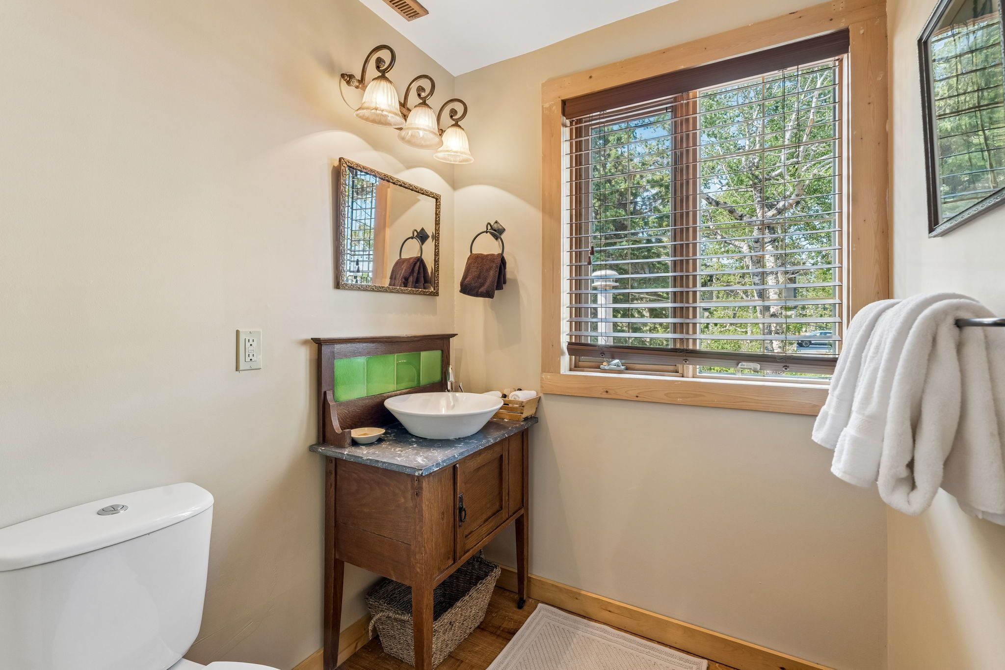 Une salle de bain comprenant des toilettes, un lavabo avec un plan de travail vert, un miroir et une fenêtre avec des stores.