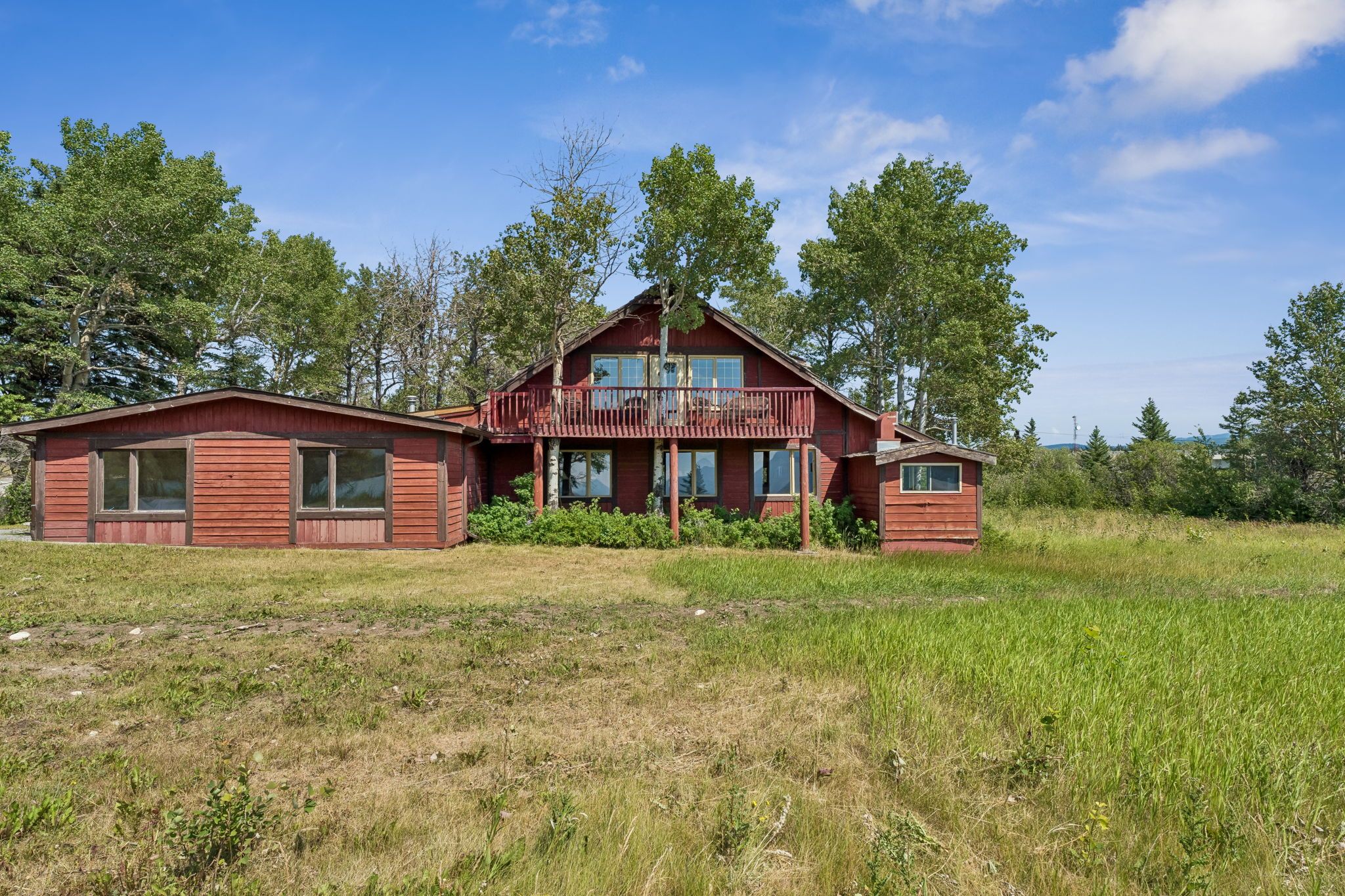 A two-story wooden cabin with a balcony, surrounded by a grassy field and trees.