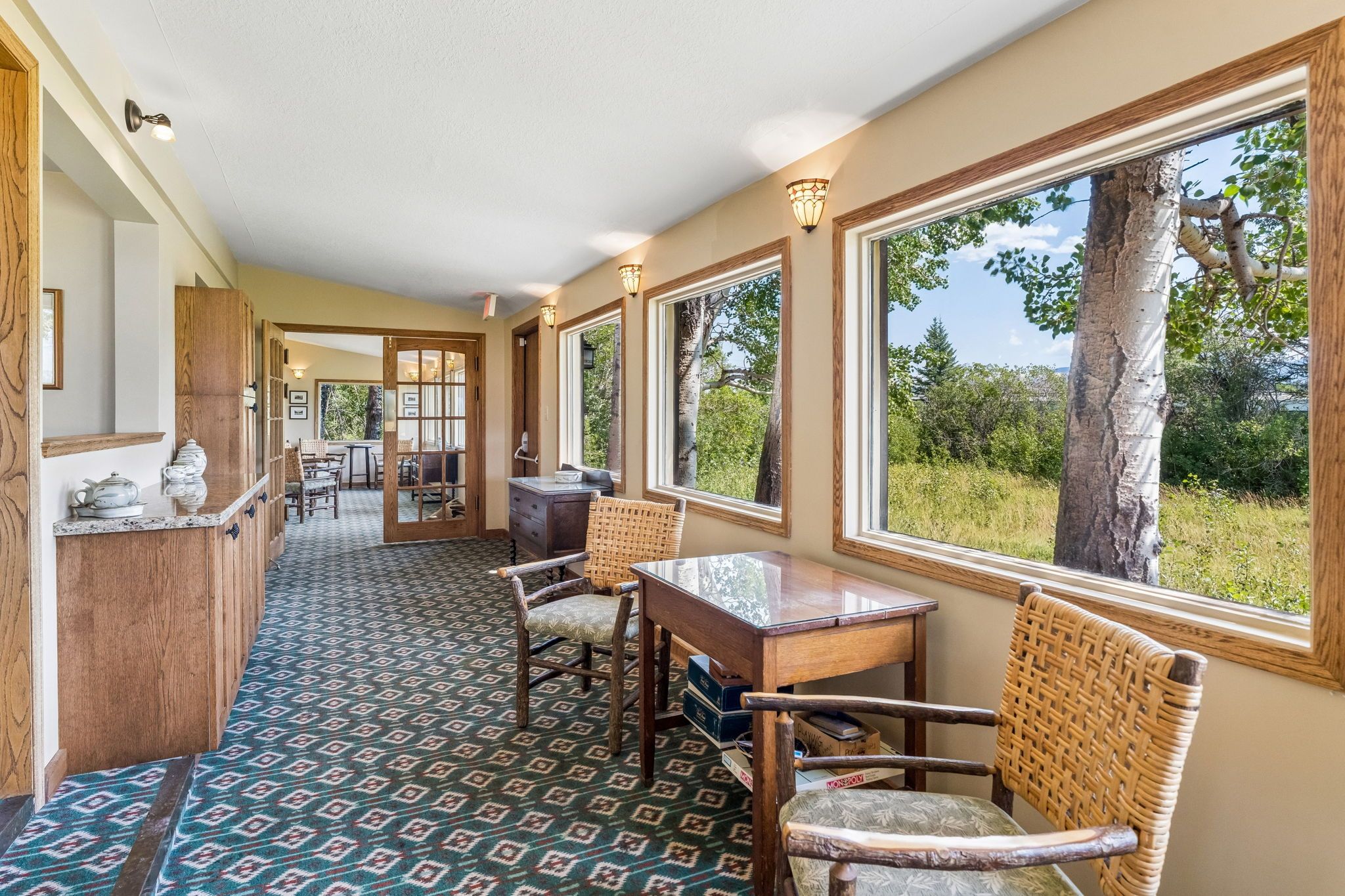A hallway with a patterned carpet, wooden furniture, and large windows offering a view of trees and sky.