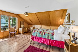 A bedroom with a wooden ceiling, a bed with patterned bedding, a nightstand with a lamp, and a window with a view of trees. - Brewsters Ranch by Canmore Premier (Seebe)