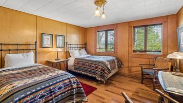A room with two beds, a desk, and a chair. There are framed pictures on the wall, a window with blinds, and a ceiling light fixture.
