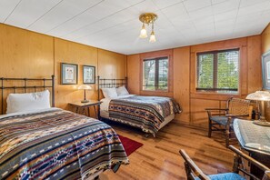 A room with two beds, a desk, and a chair. There are framed pictures on the wall, a window with blinds, and a ceiling light fixture.