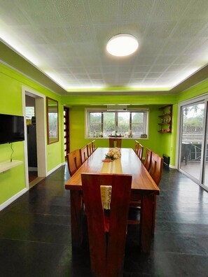 A dining area with a long wooden table, chairs, a ceiling light, and a view through large windows.