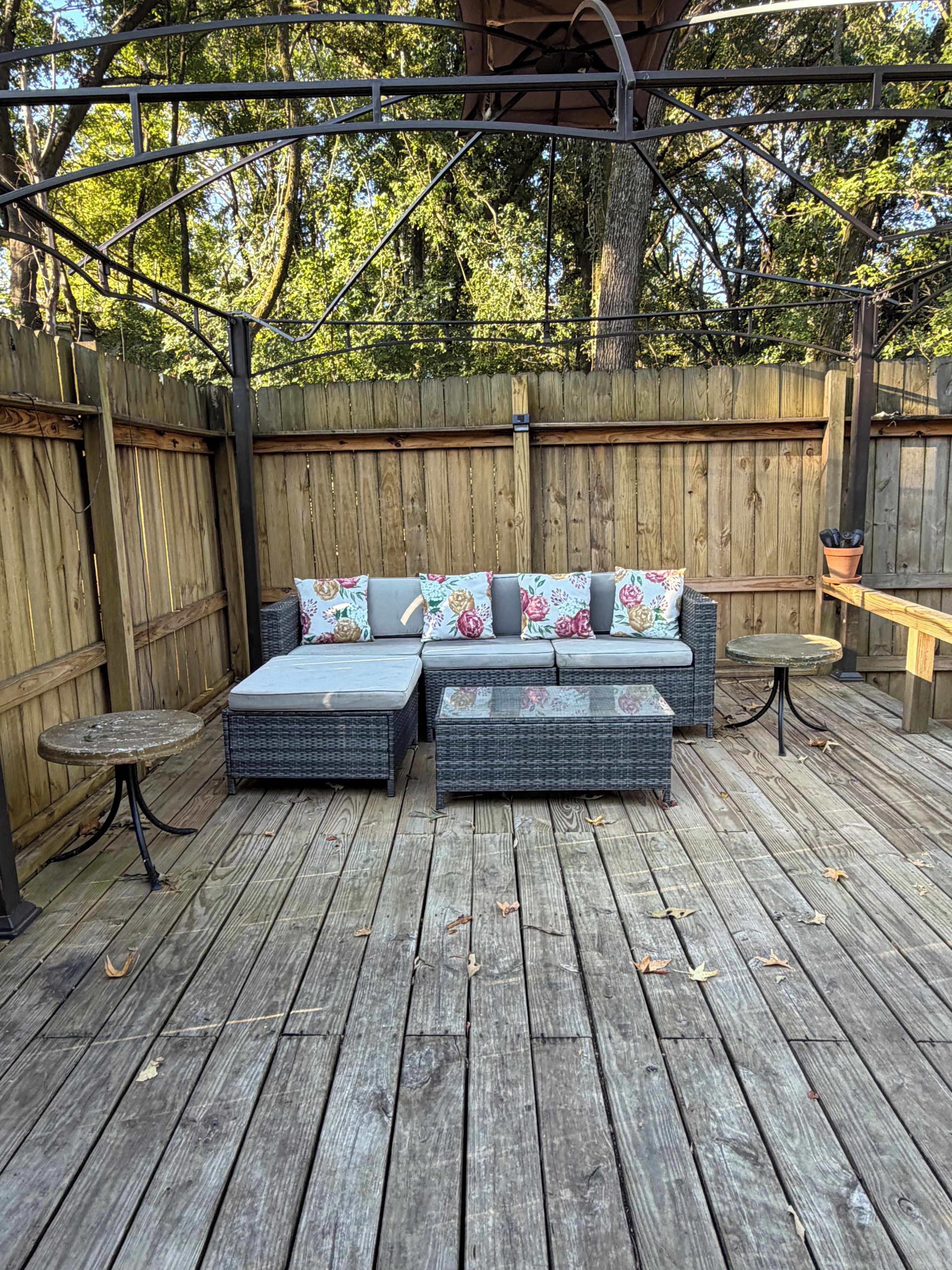 An outdoor patio with a wooden deck, a wicker sofa with floral cushions, and a small round table.