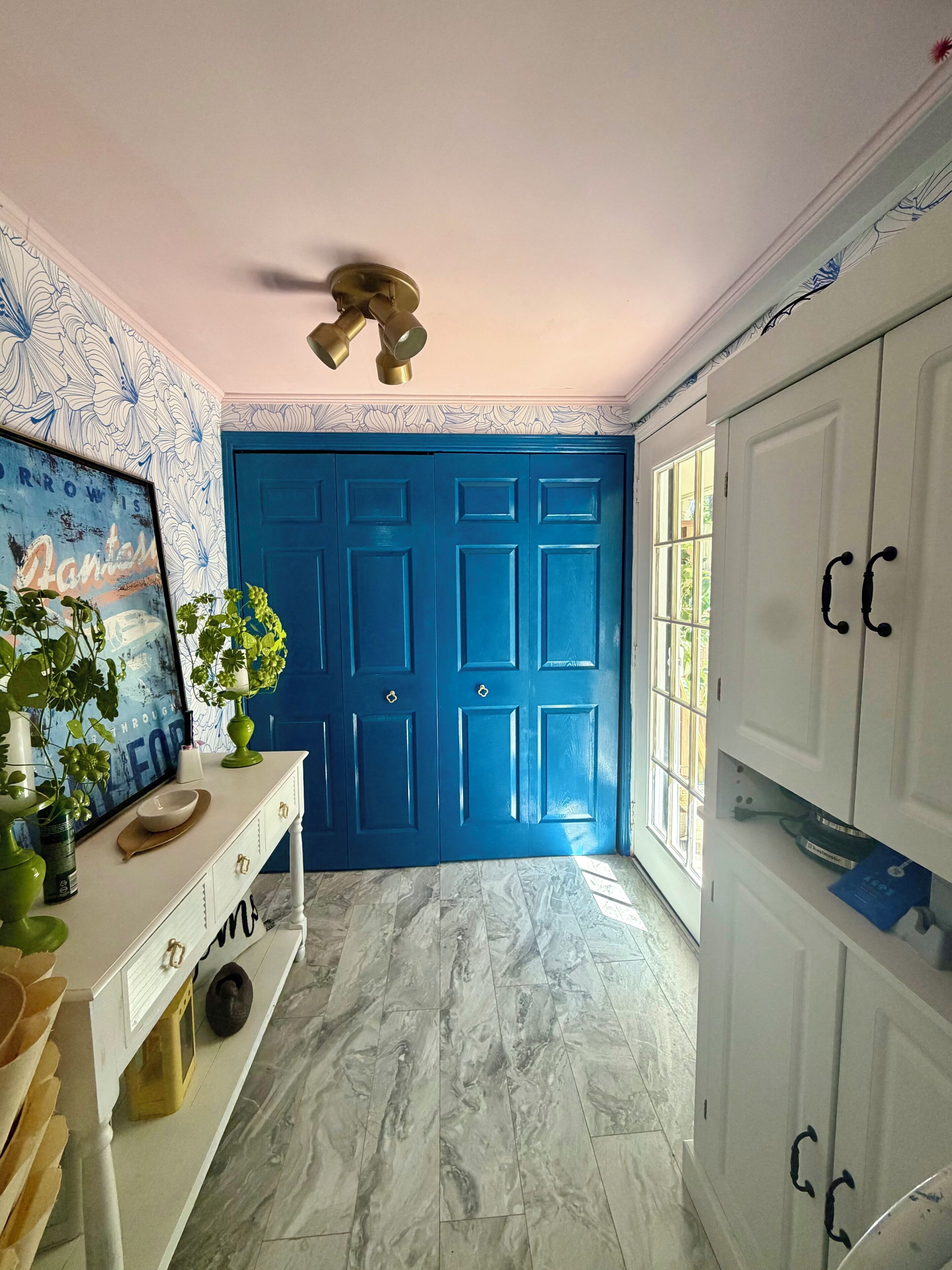Um corredor com uma porta azul, um armário branco com puxadores pretos, chão de mármore e uma parede com padrão floral.
