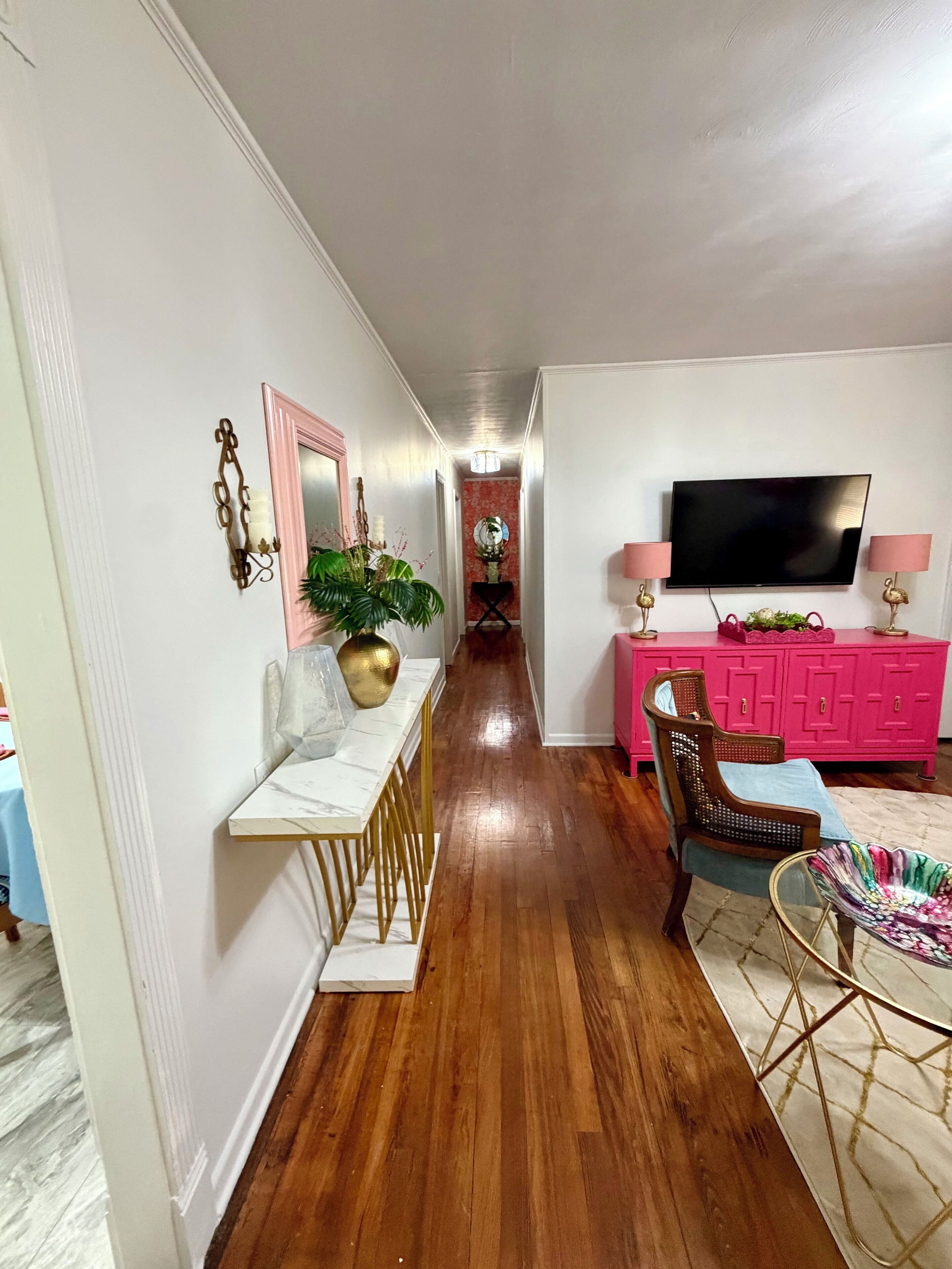 A hallway with a dining table, chairs, a pink cabinet, a television, and a plant.