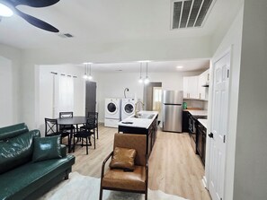 A compact living space with a dining area, a sofa, a chair, a washing machine, a refrigerator, and a kitchen with cabinets and a countertop.