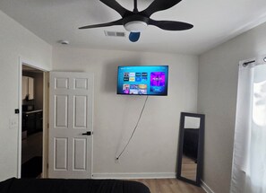 A room with a ceiling fan, a mounted flat-screen TV displaying various streaming services, a door leading to a kitchen, and a mirror on the wall.