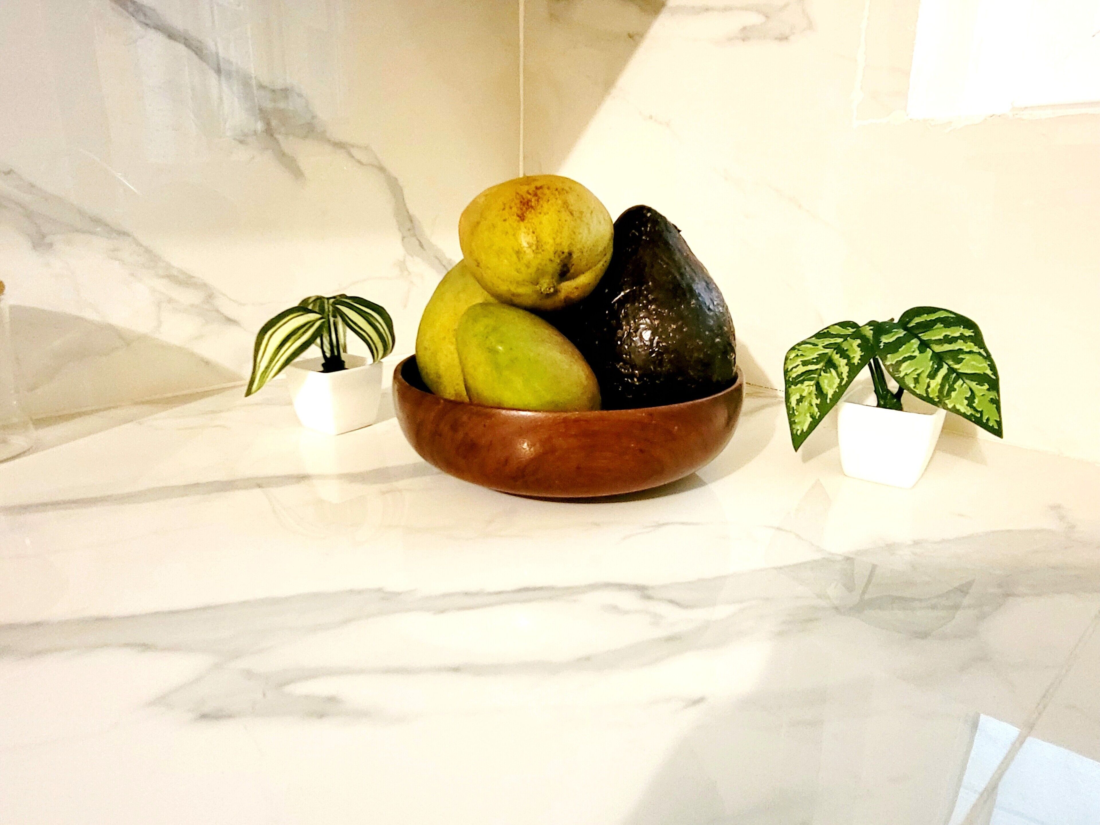 A marble countertop with a wooden bowl containing fruits and two potted plants.