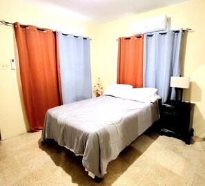 A bedroom with a bed, curtains, a nightstand, and an air conditioning unit. - Cozy 1-bedroom house with AC in Kingston neighborhood (Kingston)