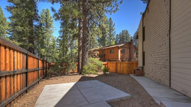 A fenced backyard with a wooden deck, a concrete patio, and a building with a brick exterior. - Summit Après | 2-Min Walk to Snow Summit (Big Bear Lake)